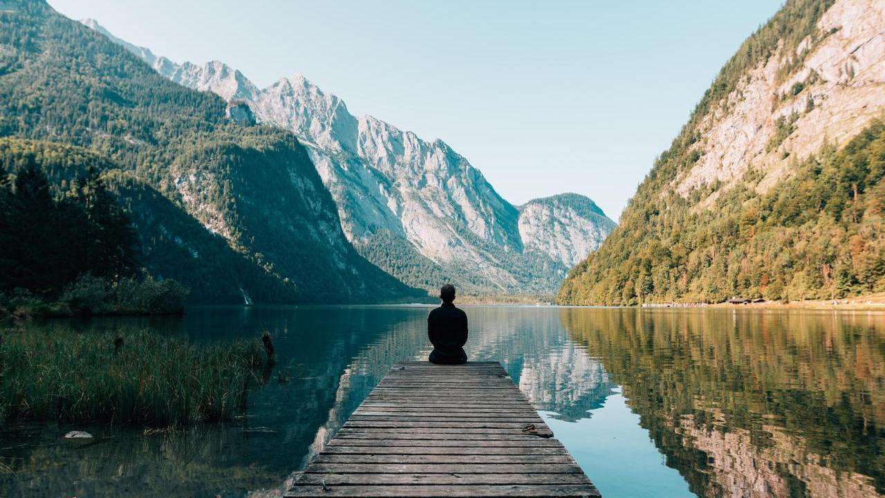 Person sitting on jetty by lake with mountains in the background